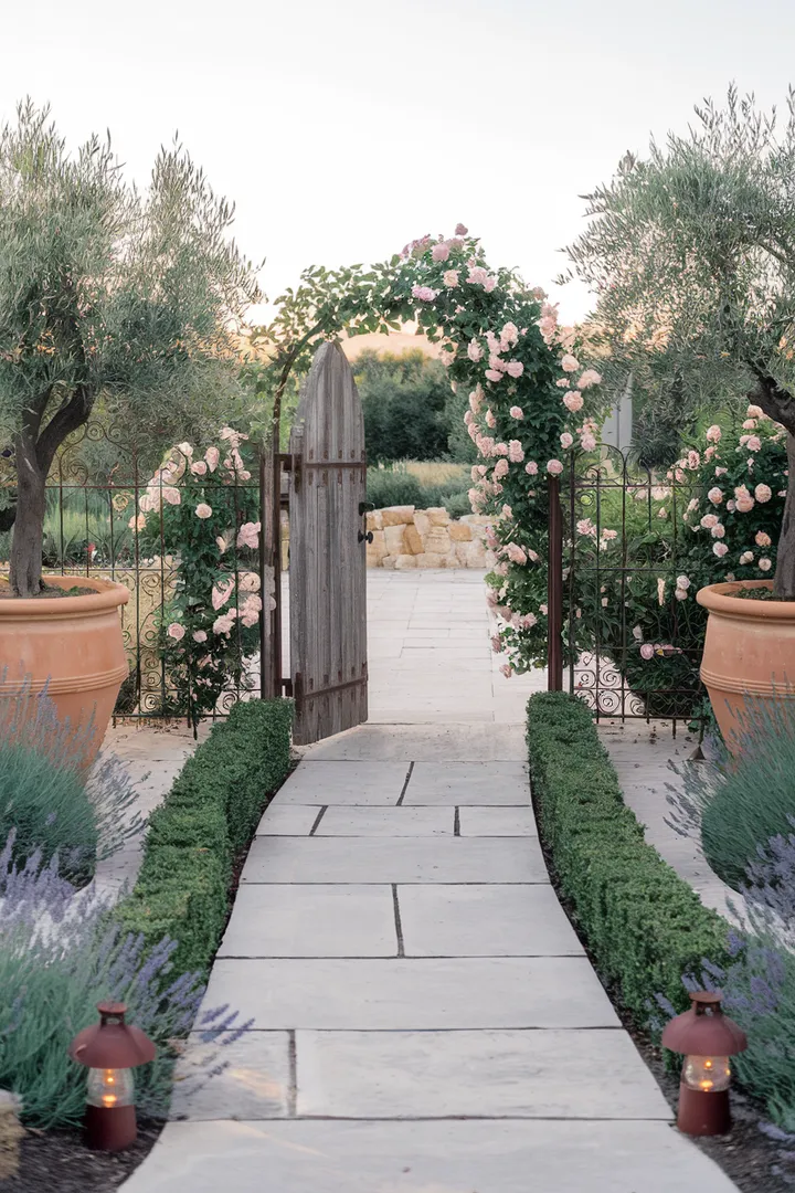 Ein romantischer Garteneingang mit einer alten Holztür, eingerahmt von duftenden Rosenranken und flankiert von Lavendel und Olivenbäumen.