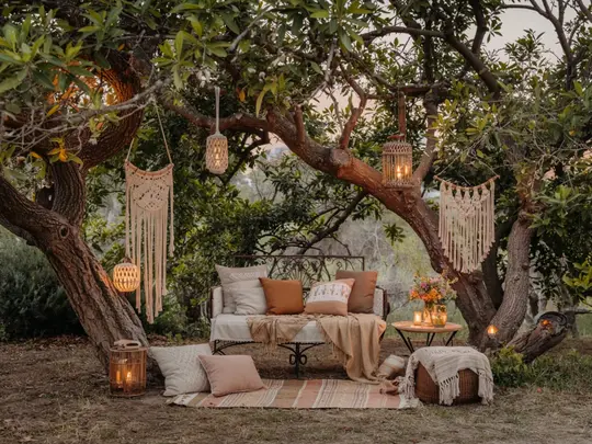 Stimmungsvoller Boho-Sitzplatz im Garten unter Bäumen – mit Vintage-Möbeln, Kissen, Makramee und warmem Abendlicht. Perfekt für romantische Gartenideen.
