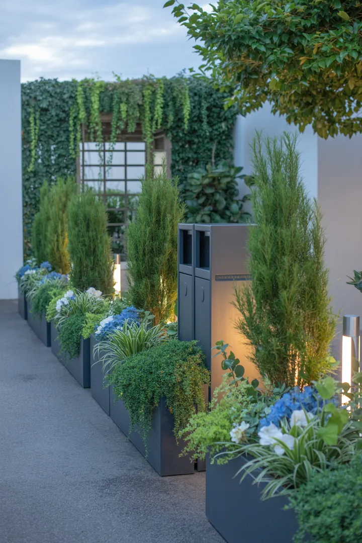 Elegante Gestaltung mit schmalen Pflanzkübeln, einem stilvollen Rankgitter, dekorativen Sichtschutzmatten, robusten Trittplatten, einer praktischen Mülltonnenbox und einer modernen Außenleuchte für den Garten.