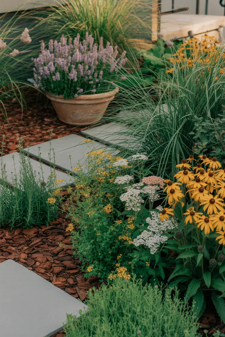 "Gestaltete Gartenlandschaft mit eleganter Beeteinfassung aus Randsteinen, umgeben von üppigen Gräsern, bunten Stauden und dekorativen Pflanzkübeln, während Unkrautvlies für Ordnung sorgt."